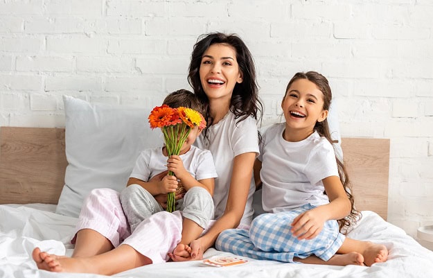 The Best 110 Happy Mother’s Day Quotes and Wishes for Moms to Show Love A happy mother with her two smiling children in bed and a bouquet of flowers.